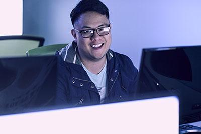 A UWF student smiles while using a computer in a dimly lit classroom.