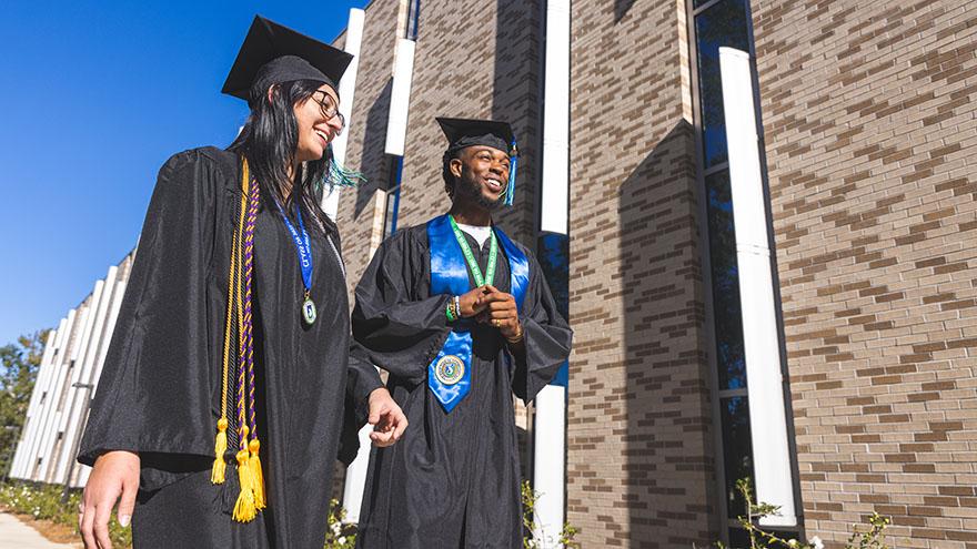 Two students stand in graduation regalia in front of an academic building.